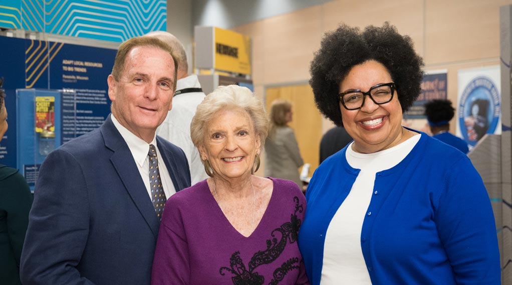 group of VIPs smile for a photo in front of the exhibit which features different multicolor informational panels