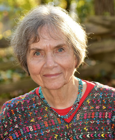 Portrait of an elderly woman with short hair, wearing a colorful embroidered top and a turquoise necklace, smiling softly against a natural background.