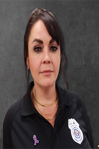 A portrait of a woman in a uniform shirt with a public safety logo, wearing necklaces and a purple ribbon pin.