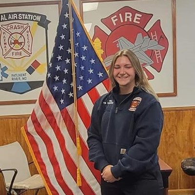 EMS graduate Paige Hill is dressed in a formal firefighter's uniform  in front of her fire department's crest and an American flag