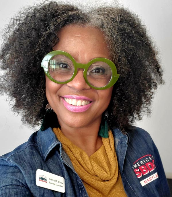 A smiling woman with curly hair and distinctive green glasses, wearing a denim jacket and a yellow sweater, representing the SBDC of Maryland.