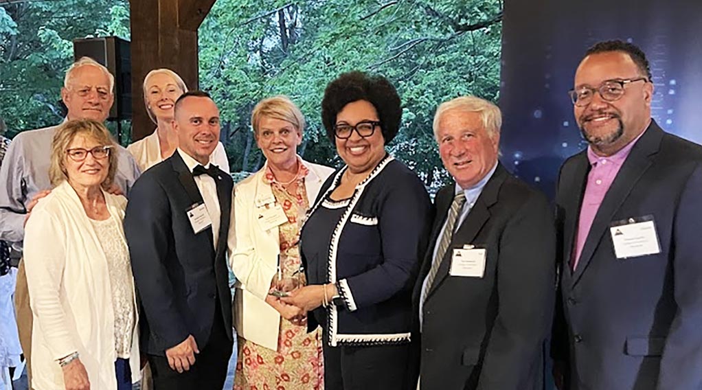 Group photo of award recipients and attendees at an event, featuring a woman holding a trophy surrounded by men and women in formal attire.