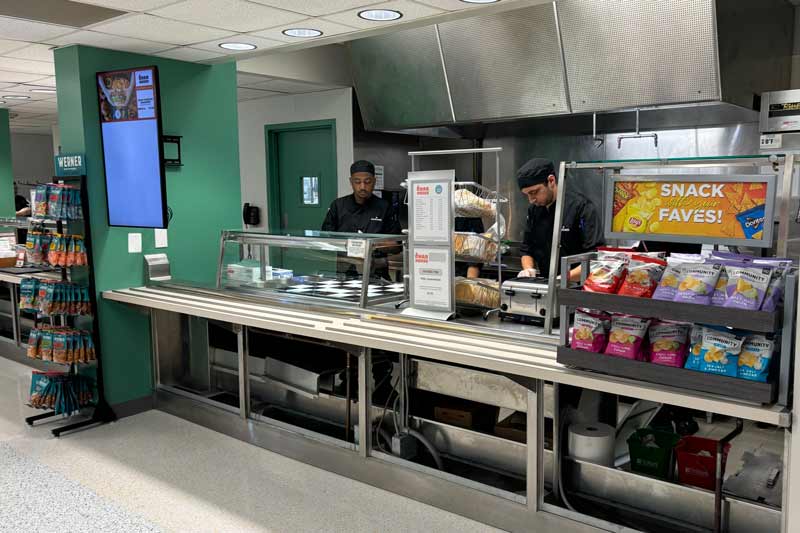 Image of a cafeteria serving area with two food workers preparing meals, accompanied by a display of snacks on the left side.